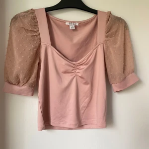 Rosa topp med puffärmar från Amisu - Säljer en ljusrosa topp från Amisu med puffiga, halvtransparenta ärmar i mesh med små broderade prickar. Toppen har en söt rynkdetalj framtill och fyrkantig ringning. Perfekt för dig som vill ha något gulligt och trendigt!