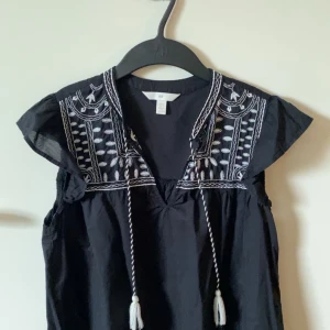 Svart blus med broderi från H&M - Svart blus från H&M med vita broderade detaljer och korta volangärmar. Blusen har v-ringning med knytband och dekorativa tofsar. Perfekt för en bohemisk look.