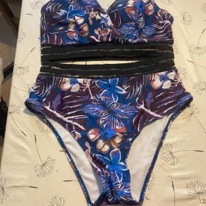 Blommig bikini med meshdetaljer - Snygg bikini med blommigt mönster i blått, lila och vitt. Överdelen har meshdetaljer under bysten och hög midja på underdelen. Perfekt för strand eller poolhäng.