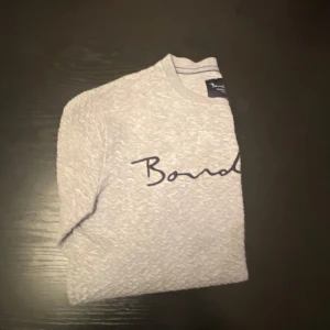 Grå sweatshirt från Bondelid - Säljer en ljusgrå sweatshirt från Bondelid med broderad logga i mörkblått över bröstet. Tröjan har rund halsringning och långa ärmar, perfekt för en avslappnad stil.