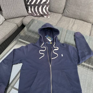 Marinblå hoodie med dragkedja från Polo Ralph Lauren - Mörkblå hoodie med vit dragsko och dragkedja från Polo Ralph Lauren. Klassisk logga broderad på bröstet och två fickor framtill. Perfekt för en avslappnad stil. Köpt från Care of Carl tror jag. Nypris runt 2000