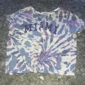Tie-dye t-shirt med MIAMI-tryck från H&M - Säljer en kortärmad tie-dye t-shirt från H&M med lila och blå nyanser och MIAMI-tryck framtill. T-shirten har knytning nertill för en cool look. Perfekt för dig som gillar färg och statement-plagg!