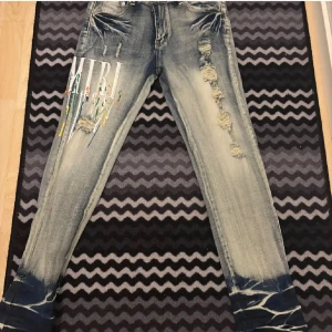 AMIRI jeans med färgstänk och slitningar - Säljer ett par snygga blå jeans från AMIRI med slitningar, färgstänk och tryckt logga på ena benet. Jeansen har klassisk femficksdesign, normal passform och unika detaljer vid bensluten. Perfekta för dig som vill sticka ut.