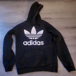 Svart hoodie från Adidas med stor logga - Svart hoodie från Adidas med stor vit trefoil-logga och text på bröstet. Klassisk känguruficka framtill 