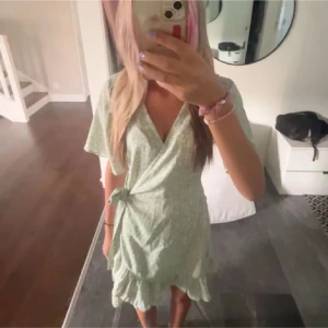 Ljusgrön omlottklänning med volang - Säljer en söt ljusgrön omlottklänning med volangkant nertill och knyt i midjan. Klänningen har korta ärmar och ett diskret blommigt mönster. Perfekt för sommarens alla tillfällen!