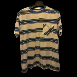 Randig t-shirt med blå detaljer - Säljer en vit t-shirt med blå, gröna och svarta horisontella ränder. T-shirten har en bröstficka med samma mönster och blå ribbad krage. Perfekt för en avslappnad stil.