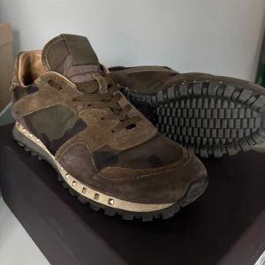 Valentino Rockrunner sneakers i brun mocka och camouflage - Säljer ett par Valentino Rockrunner sneakers i brun mocka med camouflage-mönster och ikoniska nitar runt sulan. Skorna har snörning och en robust, mönstrad sula. Perfekta för dig som gillar streetstyle med lyxig känsla. No Og, skick 8/10 (helt ärligt inga problem mer än att dem är använda, alla nitar kvar o så). Perfekta hela året om!