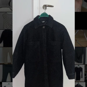 Svart teddyjacka med krage - Mysig svart teddyjacka med klassisk krage och stora framfickor. Jackan har lång ärm och stängs med knappar framtill. Perfekt för kyligare dagar och ger en trendig look.