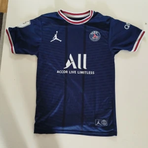 PSG fotbollströja Mbappé 7 - Mörkblå Paris Saint-Germain fotbollströja med Mbappé 7 på ryggen. Tröjan har vita och röda detaljer vid krage och ärmslut, samt klubbmärke och sponsortryck på bröstet. Tillverkad i lätt och ventilerande material.