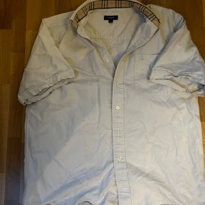 Burberry Skjorta Vit-Beige - Säljer en klassisk beige kortärmad skjorta från Burberry med den ikoniska rutiga detaljen på insidan av kragen och broderad logga på bröstfickan. Perfekt för en stilren och avslappnad look till sommarens varma dagar! Sitter relativt stort