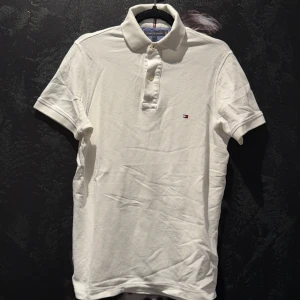 Vit piké t-shirt från Tommy Hilfiger - Klassisk vit piké t-shirt från Tommy Hilfiger med liten logga på bröstet och krage med knappar. Perfekt för en clean och stilren look.