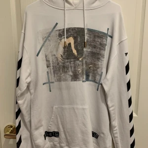 Vit hoodie från Off-White | ev byte - Säljer en vit hoodie från Off-White med stort grafiskt tryck framtill och ikoniska svarta ränder på ärmar och rygg. Tröjan har huva med dragsko och en stor magficka. Perfekt för dig som gillar streetwear och vill sticka ut.