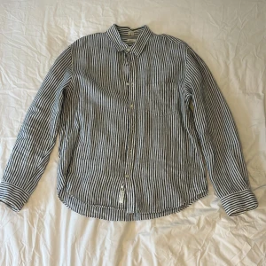 Blåvit randig skjorta Regular Fit från arket  - Snygg blå och vit randig skjorta från Arket i 100% linne. Klassisk krage, långärmad med knappar framtill och på ärmsluten. Perfekt för en clean och stilren look. Inga defekter, som ny. Passar strl M-L.