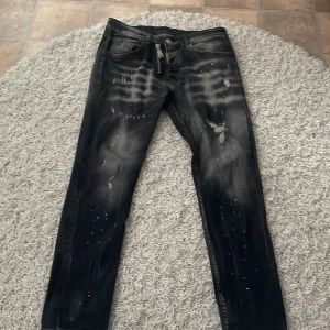 Svarta jeansbyxor från Dsquared2 med slitningar - Säljer ett par svarta Dsquared2 jeans med slitningar, vita färgstänk och dragkedjedetalj fram. Bak finns en stor vit patch med texten 'ICON'. Snygg streetstil och raka ben.