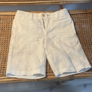 Vita linneshorts från Polo Ralph Lauren - Snygga vita shorts i linne från Polo Ralph Lauren med klassisk passform. De har dragkedja och knapp framtill, bälteshällor och två bakfickor med knappar. Perfekta för varma dagar och en stilren look.Cirka storlek 150cm.