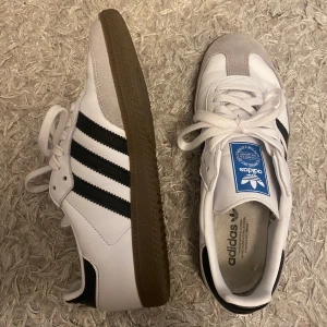 Adidas Samba sneakers  - Säljer ett par klassiska Adidas Samba sneakers i vitt med svarta ränder och grå mockadetaljer vid tån. Skorna har vit ovandel, bruna gummisulor och snörning. Köpta för bara någon månad sedan så fortfarande väldigt fint skick! Kartong följer med.