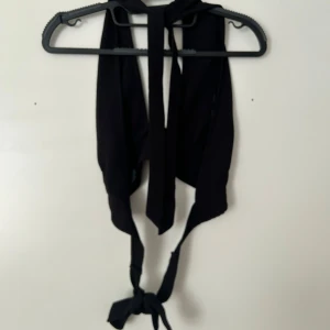 Svart omlott topp med knyt - Säljer en svart omlott topp med knyt framtill. Toppen är ärmlös och har en djup v-ringning. Perfekt för att styla med höga jeans eller kjol. Snygg och trendig modell som ger en cool look.