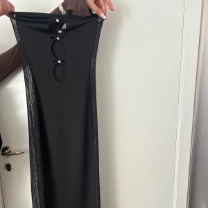 Svart långklänning med cut-outs - Säljer en svart långklänning med snygga cut-outs framtill och dekorativa pärldetaljer. Klänningen har en tight passform och är ärmlös, perfekt för dig som vill sticka ut på festen.