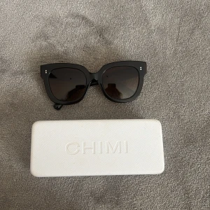 Bruna solglasögon från CHIMI - Model 08!! Snygga svarta solglasögon från CHIMI med bred båge och mörka glas. Klassisk fyrkantig form och diskreta silvriga detaljer vid fronten. Kommer med vit CHIMI-fodral.
