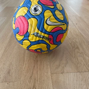 Premier League Nike Flight fotboll 21/22 - Säljer en officiell matchboll från Premier League säsongen 21/22, Nike Flight modell. Bollen har ett färgstarkt mönster i blått, gult och rött med svarta detaljer och Premier League-logga. Storlek 5 och FIFA Quality-märkt.