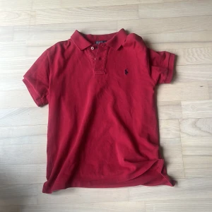 Röd pikétröja från Polo Ralph Lauren - Säljer en klassisk röd pikétröja från Polo Ralph Lauren med broderad logga på bröstet. Tröjan har korta ärmar, krage och knappar framtill. Tillverkad i 100% bomull för en skön och luftig känsla.