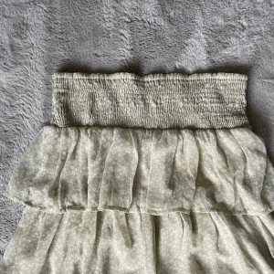 Ljusbeige volangkjol med smock - Supersöt ljusbeige kjol med bred smock i midjan och dubbla volanger. Kjolen har ett diskret blommigt mönster och är gjord i ett lätt, luftigt material som passar perfekt till varma dagar.