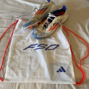 Adidas F50 fotbollsskor med väska - Säljer ett par Adidas F50 fotbollsskor i vitt med blå och röda detaljer. Skorna har snörning och platt sula med fasta dobbar, perfekt för spel på gräs. Medföljer en vit meshväska med F50-logga och röda snören.