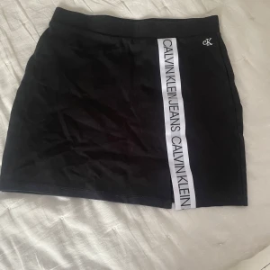 Svart kjol från Calvin Klein Jeans - Svart kjol från Calvin Klein Jeans med vit logoremsa längs framsidan. Kjolen har en tight passform och elastisk midja. Perfekt för en sportig och trendig look. Diskret CK-brodyr upptill.