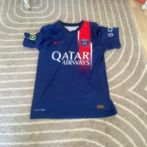 PSG fotbollströja Mbappé #7 - Säljer en blå Paris Saint-Germain fotbollströja med Mbappé #7 på ryggen. Tröjan har röd och vit rand på framsidan, Qatar Airways sponsortryck och PSG-emblem på bröstet. Korta ärmar och tillverkad i lätt funktionsmaterial. 
