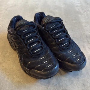 Svarta Nike Air Max Tn - Säljer ett par svarta Nike Air Max Plus sneakers med Tn-detaljer på hälen och gula accenter under sulan. Skorna har snörning, mesh- och syntetöverdel samt en chunky sula med synliga luftkuddar. Perfekta för dig som gillar streetwear och sportig stil.