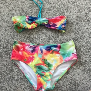 Färgglad blommig bikini - Säljer en färgglad bikini med blommigt mönster i rosa, gul, grön och blå. Överdelen är bandeau-modell med knyt i nacken och underdelen är klassisk trosa. Perfekt för stranden eller poolen!