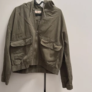 Olivgrön vindjacka från ONLY - Snygg olivgrön vindjacka från ONLY med huva, dragkedja och två stora fickor framtill. Jackan har resår i midjan och vid ärmsluten för en skön passform. Perfekt för dig som gillar en avslappnad och cool stil.