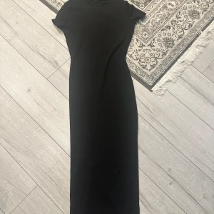 Svart långklänning med kort ärm - En stilren svart långklänning med rund hals och korta ärmar. Klänningen har en rak och enkel passform som ger en clean look. Perfekt för dig som gillar minimalistisk stil och vill ha något som funkar till många olika tillfällen.