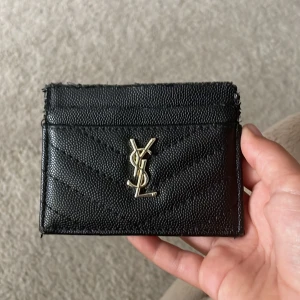 Svart YSL plånbok i skinn - Snygg svart plånbok från YSL med klassisk guldlogga framtill. Plånboken är gjord i strukturerat skinn med quiltad design och har flera fack för kort. Perfekt storlek för att ha med sig överallt och ger en lyxig känsla till vardagen. Är lite sliten på toppen men syns inte så mycket.