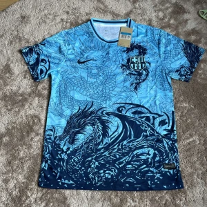 Blå Barcelona x Nike fotbollströja drake - Snygg Barcelona fotbollströja från Nike i ljusblått med mörkblå drakmotiv över hela tröjan. Klassisk rund hals med blå/vit rand, klubbmärke på bröstet och Nike-logga. Tillverkad i lätt och ventilerande funktionsmaterial, perfekt för match eller träning.