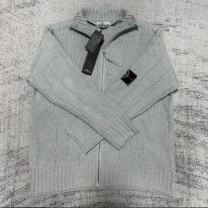 Ljusgrå stickad kofta från Stone Island - Säljer en ljusgrå stickad kofta från Stone Island med hel dragkedja och ribbad krage. Tröjan har lång ärm, diskret mönster i stickningen och den klassiska Stone Island-loggan på ärmen. Perfekt för lager-på-lager och snygg till jeans.