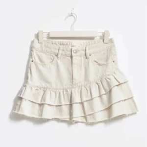 Vit volangkjol i jeans från Gina Tricot  - Supersöt vit kortkjol i jeans från Gina Tricot med volanger och råa kanter. Kjolen har klassisk femficksdesign, bälteshällor och knäppning fram. Perfekt 