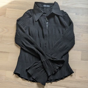 Svart plisserad skjorta  - Snygg svart skjorta  med plisserat tyg och klassisk krage. Skjortan har långa ärmar och knappar framtill. Perfekt för dig som gillar en stilren och trendig look.