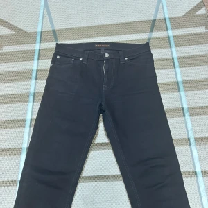 Svarta nudie jeans 29/32 - Svarta jeans från Nudie Jeans med klassisk femficksdesign och diskret broderad bakficka. Jeansen har smal passform och är tillverkade i stretchig bomull för extra komfort. Perfekta för dig som gillar stilrena och tidlösa jeans. Använda två gånger sparsamt. 29 midja 32 längd. Skriv om ni har några frågor eller funderingar så svarar jag så snabbt som möjligt
