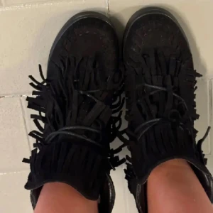 Svarta boots med fransar i mocka - Snygga svarta boots i mocka med coola fransdetaljer runt skaftet och snörning framtill. Skorna har en platt sula och ger en trendig boho-vibe, perfekta till jeans eller klänning.