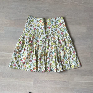 Blommig kjol från Moschino Cheap and Chic - Superfin kjol från Moschino Cheap and Chic med färgglatt blommönster i gult, rosa och blått på vit botten. Kjolen är kort och har veckad design som ger extra volym. Perfekt för dig som vill sticka ut med en lekfull och retro vibe.