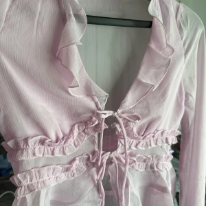 Ljusrosa volangblus med snörning - Supersöt ljusrosa blus med volangdetaljer längs urringningen och midjan. Blusen har lång ärm, omlottkänsla och snörning framtill. Tunt, lätt material som känns luftigt och fräscht. Aldrig använd 