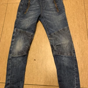Regular jeans från Lindex med förstärkta knän. Storlek 128 - Säljer ett par tåliga och bekväma jeans från Lindex, modell Regular, i färgen blå. Storlek 128, passar pojkar ca 7–8 år. Jeansen har förstärkta knän, resår i midjan och bensluten samt dragkedjefickor framtill – perfekta för aktiva barn!  Jeansen är använda men i fint skick – inga hål eller fläckar.  Perfekta för både lek och vardag! 