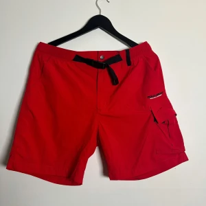 Röda Polo Sport badbyxor - Riktigt snygga röda badbyxor från Polo Sport Ralph Lauren med svart justerbart bälte och praktisk cargoficka på sidan. Shortsen har meshfoder och elastisk midja för extra komfort. Perfekta för sommardagar eller chill vid stranden.