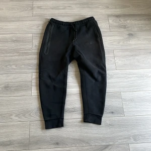 Nike tech byxor - Svarta mjukisbyxor med elastisk midja och snörning. Byxorna har smal passform vid anklarna och ribbade muddar. Perfekta för chill eller träning, med dragkedjeförsedda fickor på sidorna. Tillverkade i mjukt material för maximal komfort.