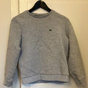 Grå sweatshirt från Lacoste - Klassisk grå sweatshirt från Lacoste med rund hals och diskret krokodillogo på bröstet. Tröjan har ribbade muddar vid ärmslut och nederkant, samt lång ärm. Perfekt basic att ha i garderoben för en clean och stilren look.