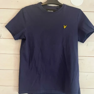 mörkblå t-shirt från Lyle & Scott - Snygg mörkblå t-shirt från Lyle & Scott med klassisk passform. T-shirten har korta ärmar och är gjord i mjuk bomull. På bröstet sitter den ikoniska gula örnloggan som ger en clean och stilren look.