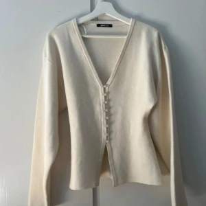 Beige kofta med v-ringning från Gina Tricot - Superfin beige kofta från Gina Tricot med djup v-ringning och dekorativa knappar framtill. Koftan har långa ärmar och är tillverkad i ett mjukt, stickat material som känns skönt mot huden. Perfekt att styla med jeans eller kostymbyxor för en clean look.