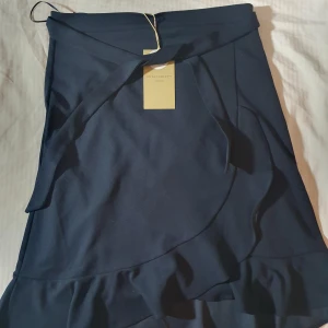 Mörkblå omlottkjol från Soyaconcept - Supersnygg mörkblå omlottkjol från Soyaconcept i återvunnen polyester. Kjolen har volangdetaljer längs nederkanten och knytband i midjan för en extra stylish look. Perfekt för dig som gillar en feminin och trendig stil.