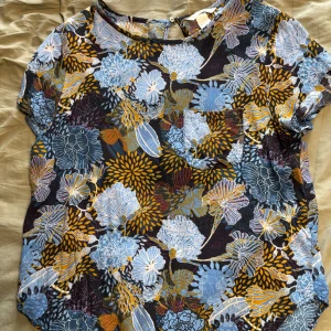 Blommig blus från H&M i storlek 42 - Färgglad blus från H&M med stort blommönster i blått, gult och lila på svart botten. Modellen har korta ärmar, rund halsringning och knapp i nacken. Tunt och luftigt material, perfekt för dig som gillar statement-plagg.